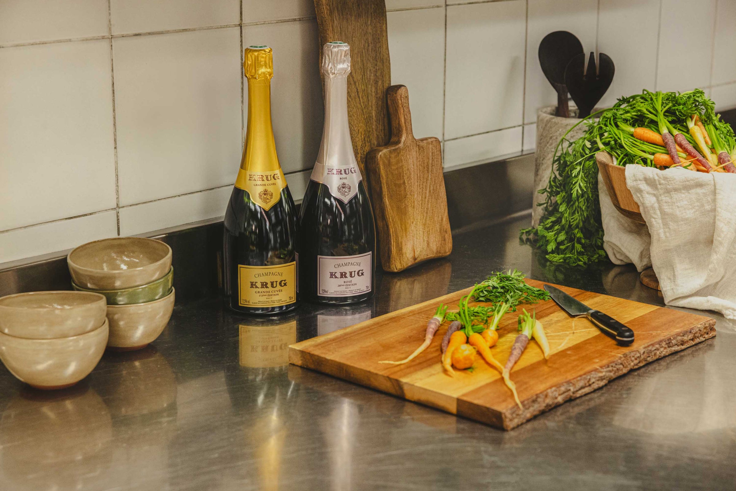 To Krug champagneflasker, Grand Cuvée og Rosé, med friske gulerødder og skål.