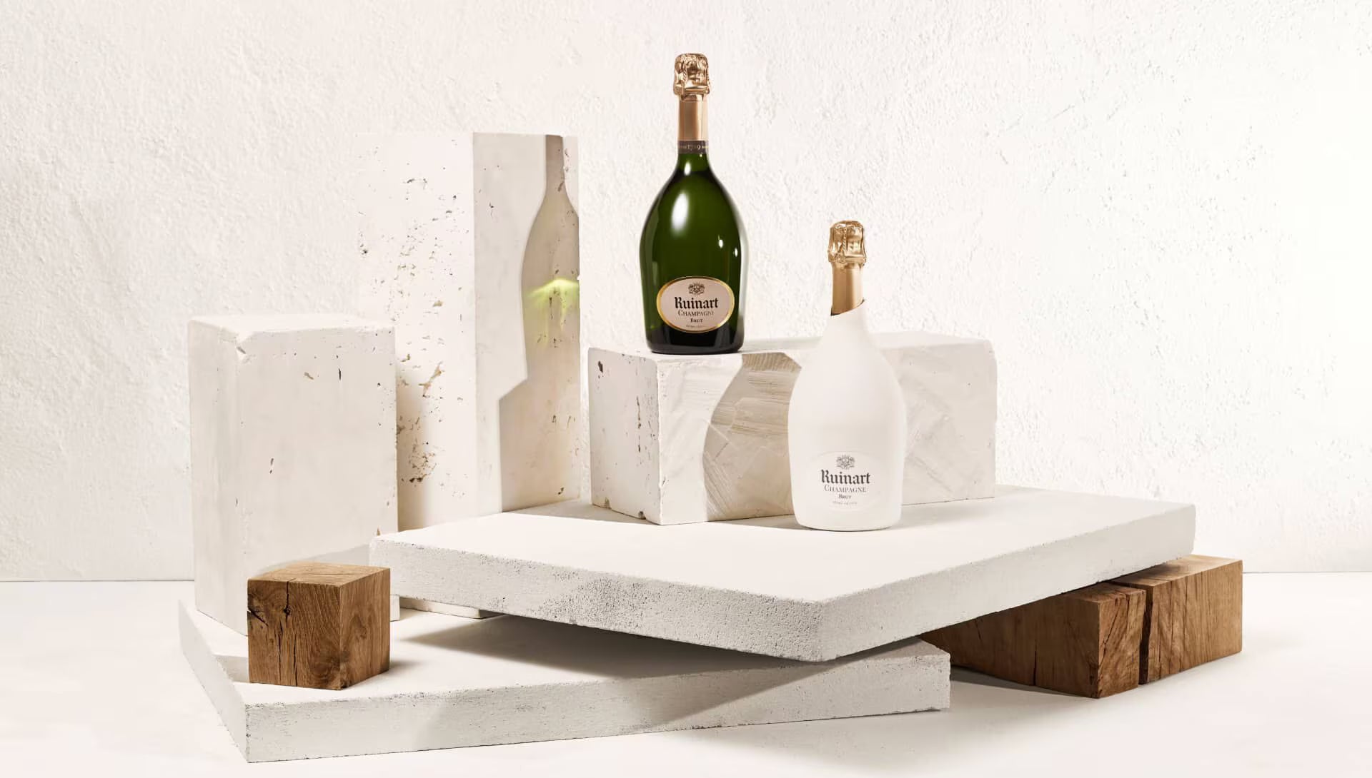 To Ruinart Brut champagneflasker på hvide og træpodier mod hvid baggrund