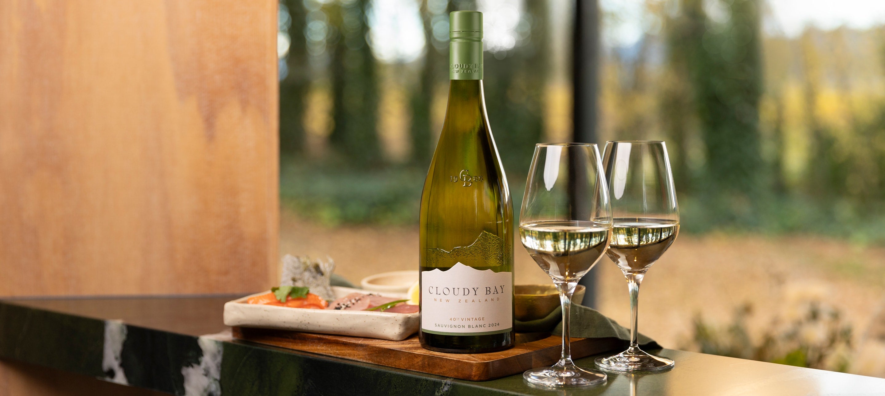 Flaske Cloudy Bay Sauvignon Blanc 2024 og to vinglas med vin, sushi i baggrunden.