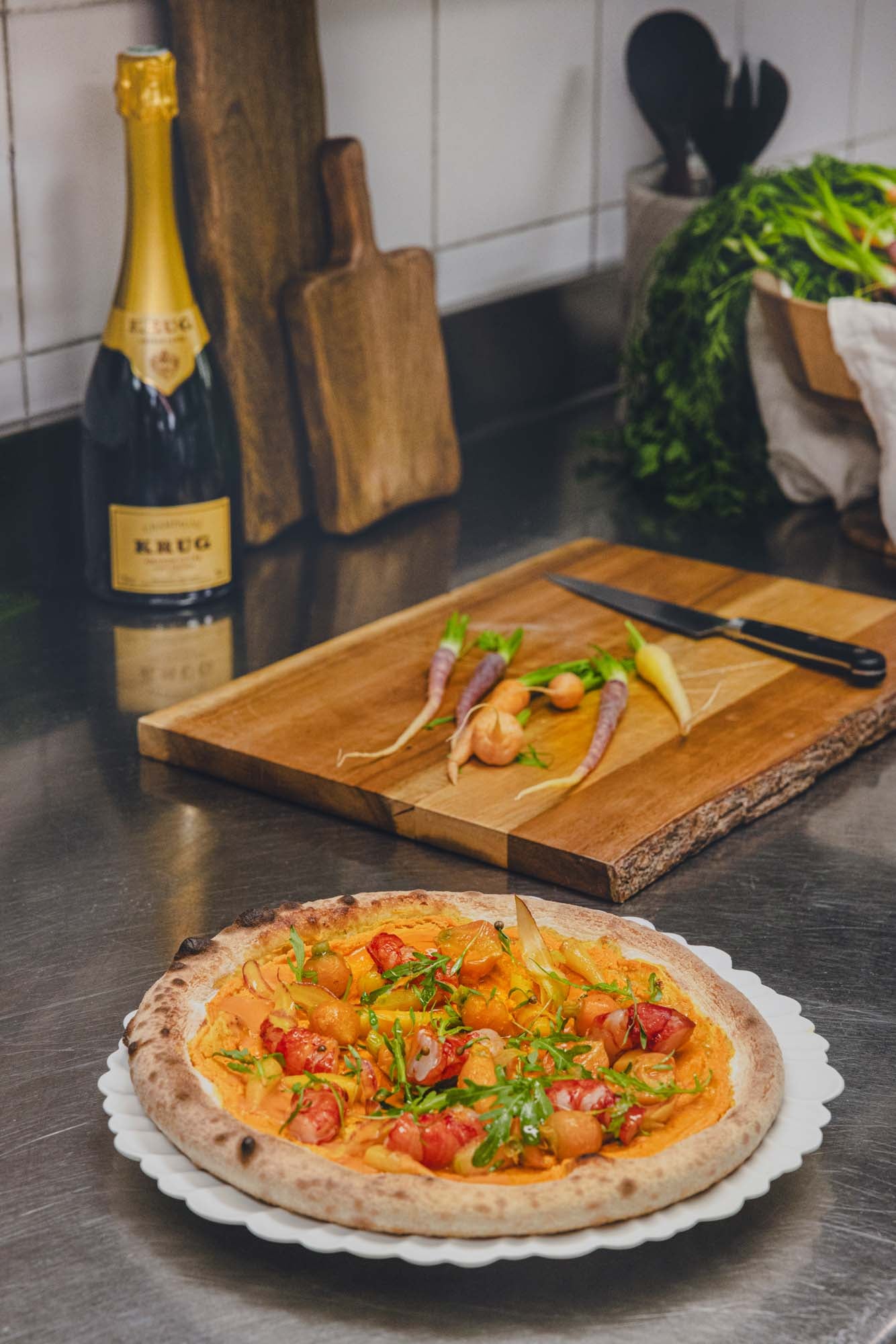 Champagneflaske, gulerødder, pizza med grøntsager, og skærebræt på køkkenbord.