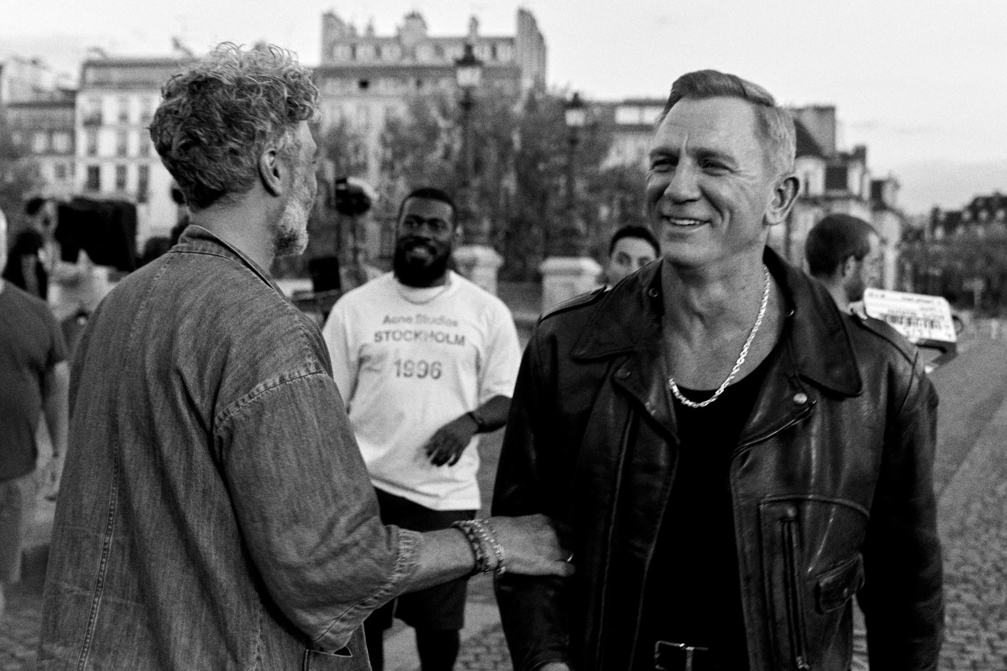 Daniel Craig i sort læderjakke og sølvhalskæde, smilende på gaden