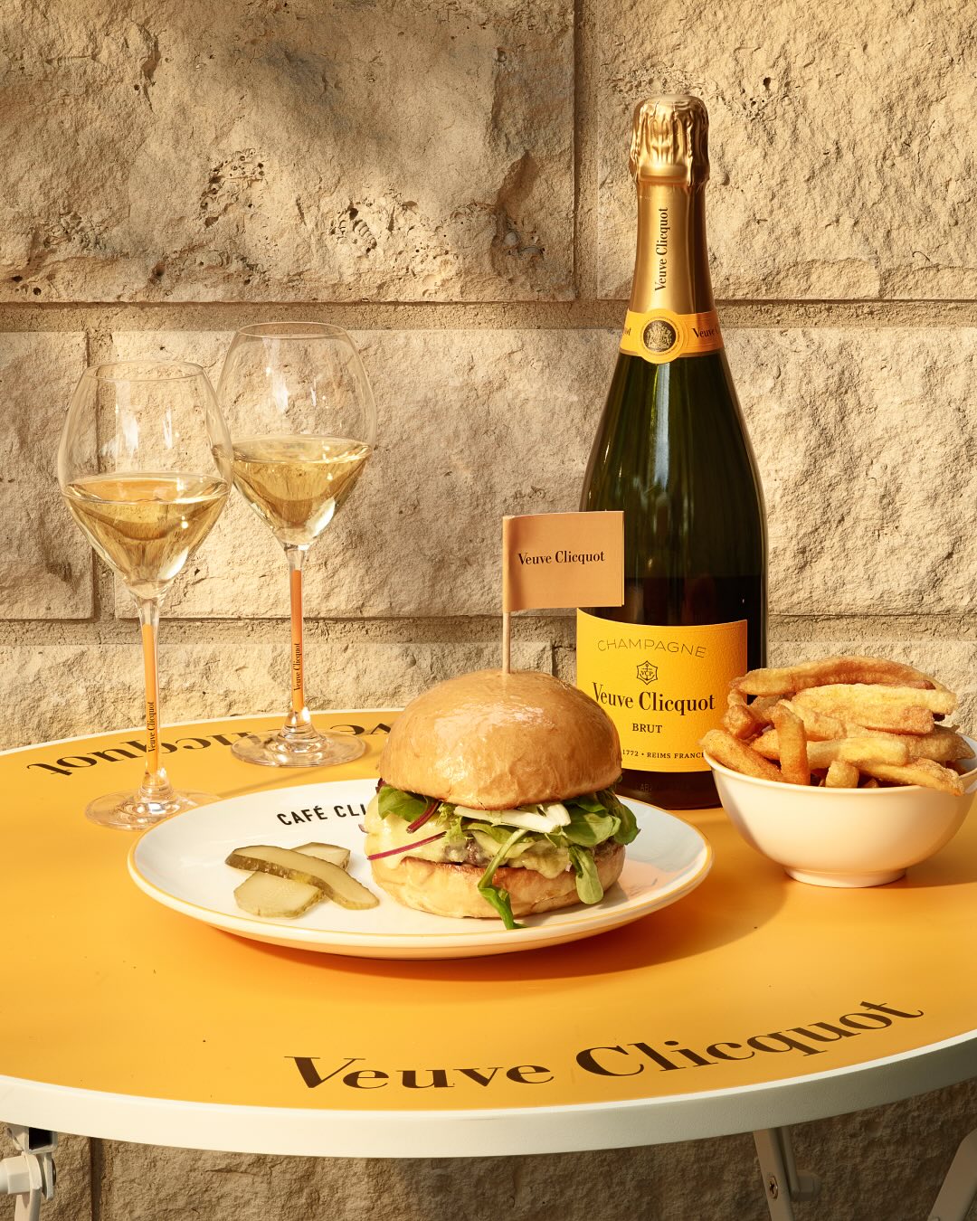 Veuve Clicquot champagneflaske, burger, pommes frites på gult bord med vinglas
