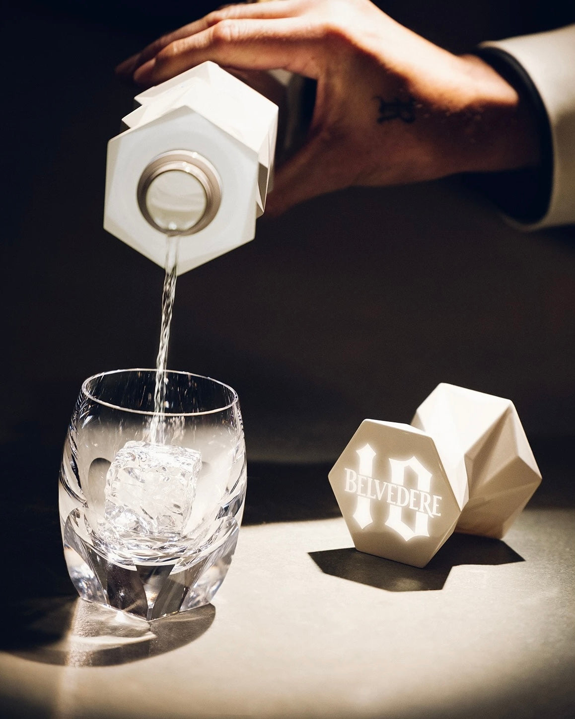 Hånd hælder Belvedere 10 vodka fra flaske i glas med isterning.