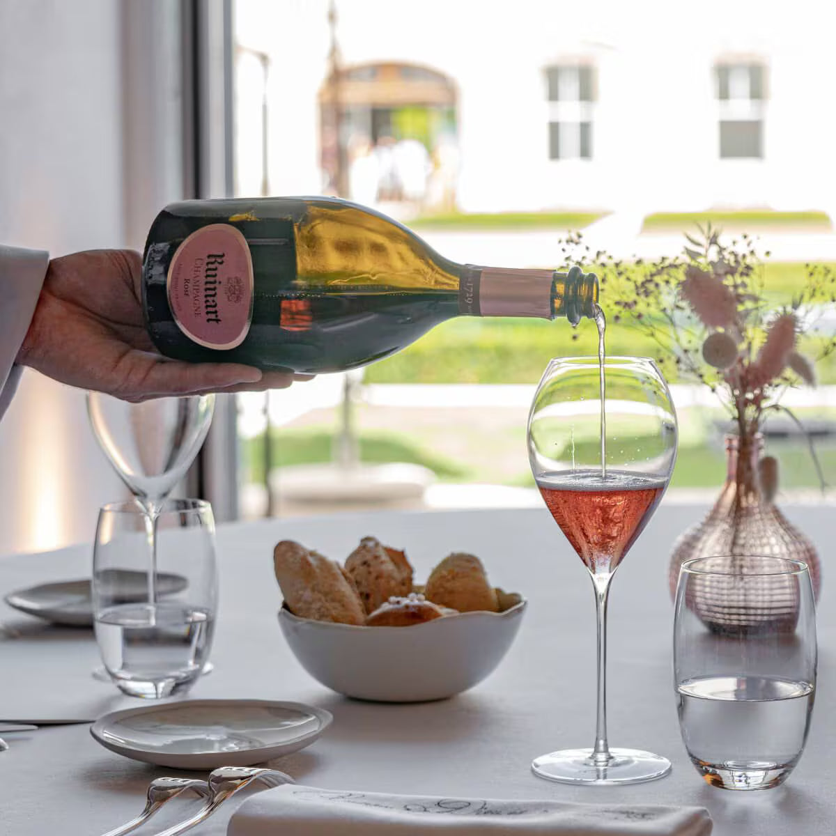 Hånd hælder Ruinart Rosé Champagne fra flaske i champagneglas på bord