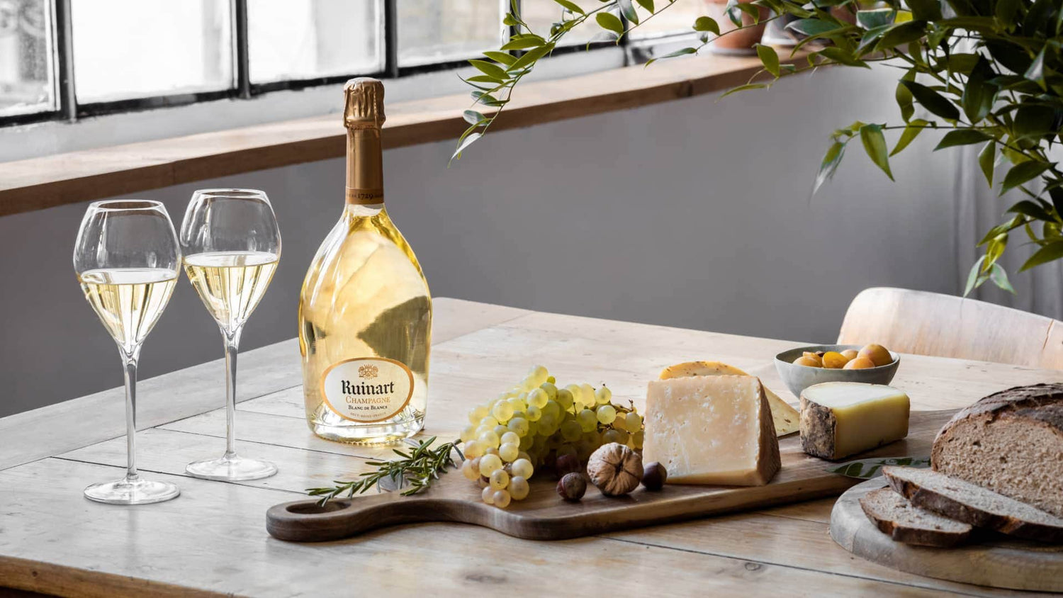 Ruinart Blanc de Blancs champagne, glas, druer, ost og brød på et træbord
