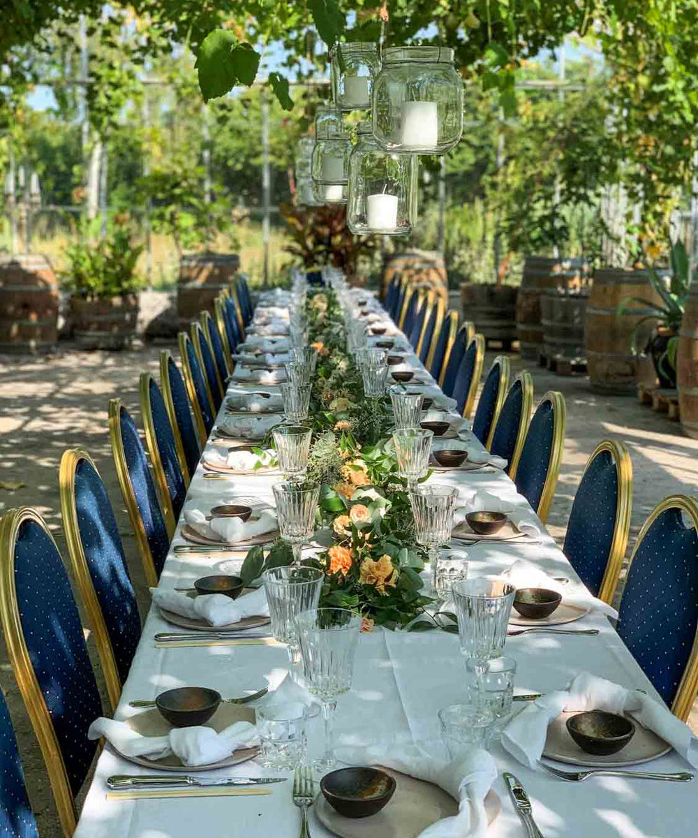 Festdækket bord med hvide duge, blå stole og stearinlyslygter hængende fra vinranker