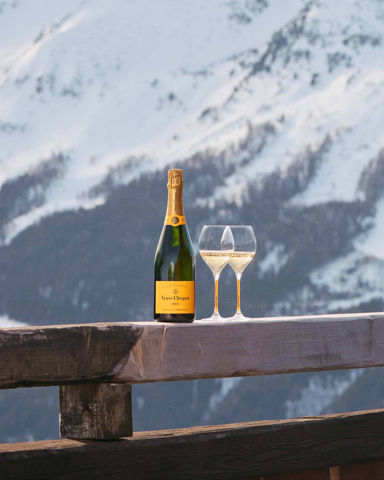 Veuve Clicquot champagne og to glas på udendørs træbord med sneklædte bjerge i baggrunden